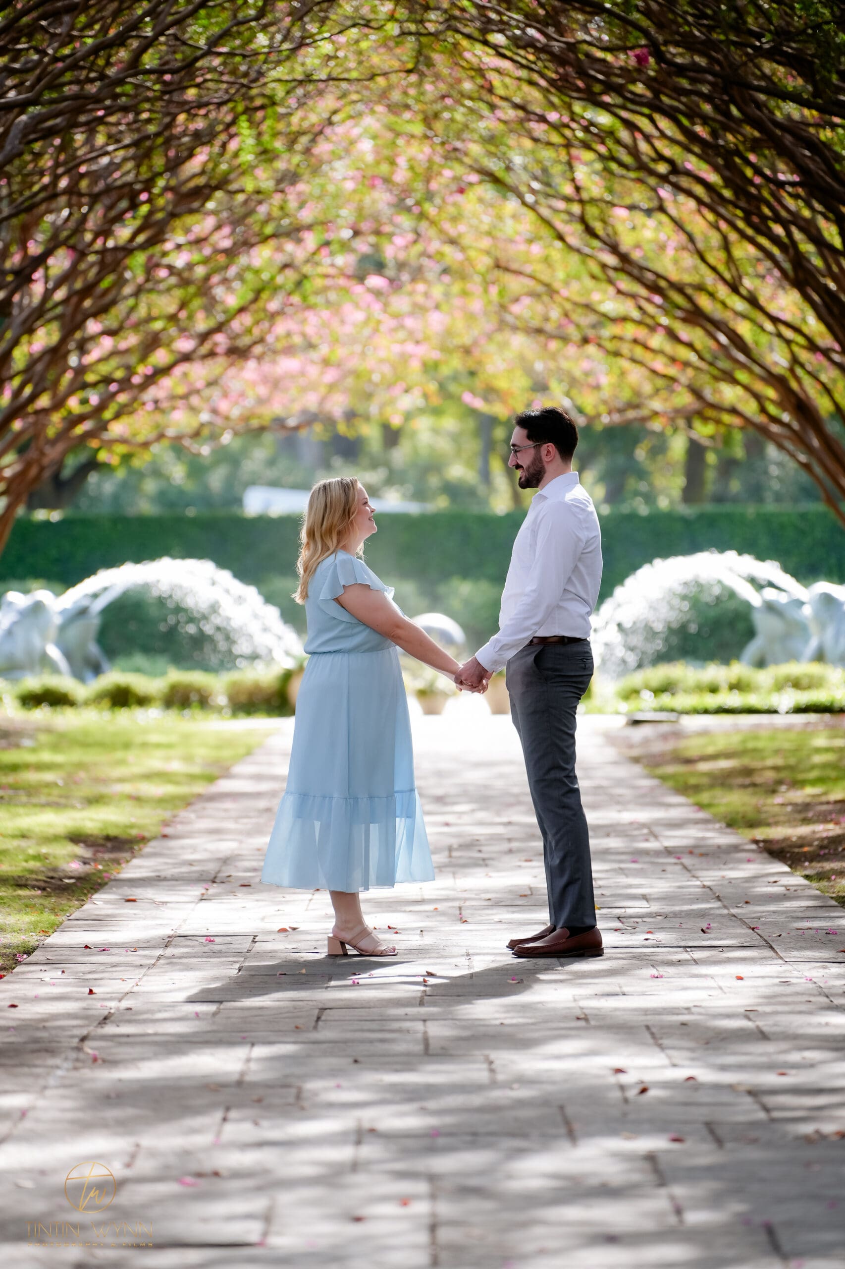 A couple faces each other and holds hands beneath arching trees, with fountains behind them.