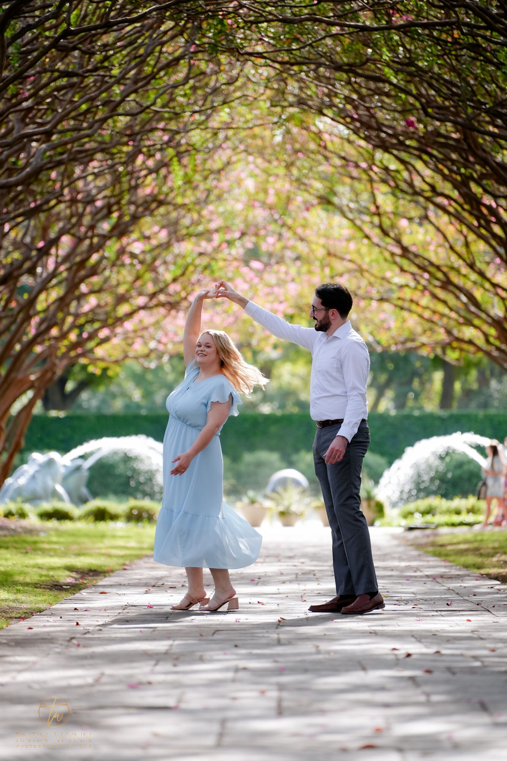 A couple twirls with hands joined overhead on a tree-lined path in front of fountains.