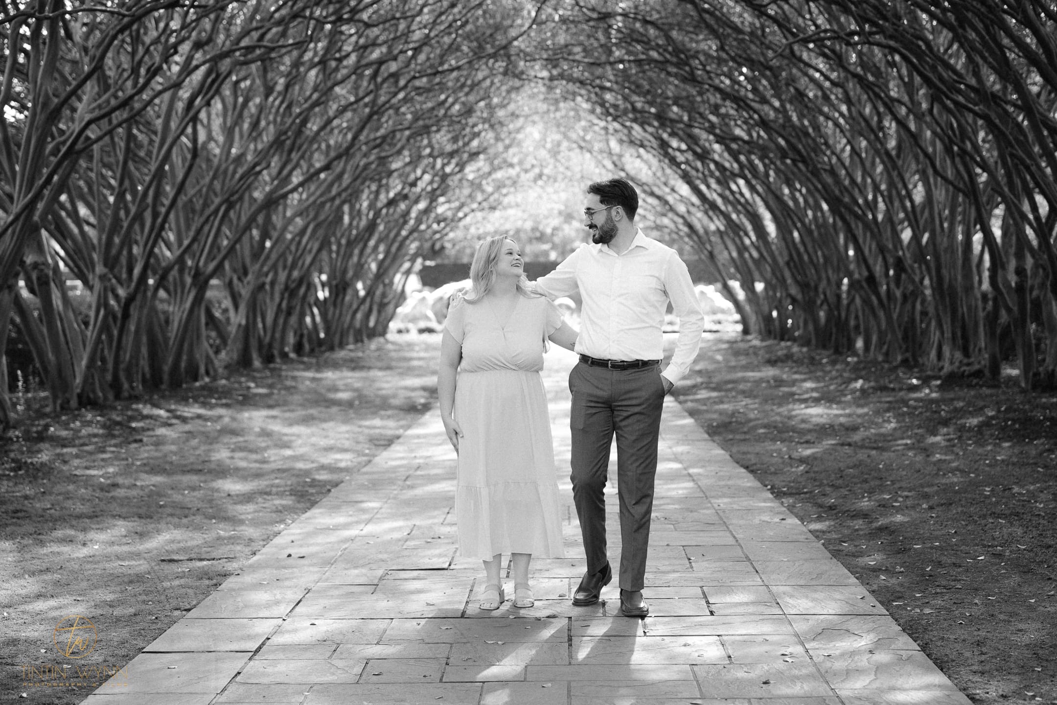 Black-and-white photo of a couple walking arm in arm beneath arching trees.