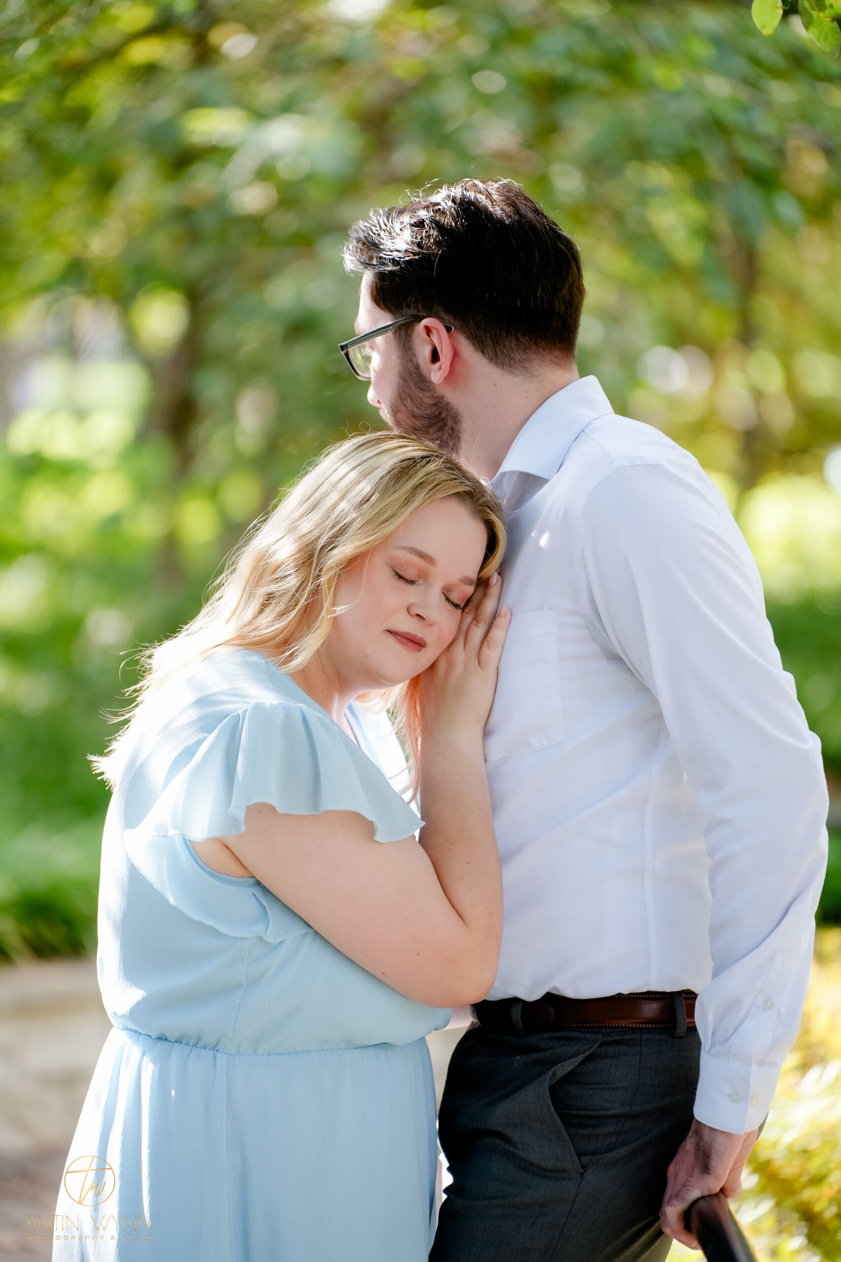 One partner rests their head against the other's chest beside a railing, with soft green foliage behind them.