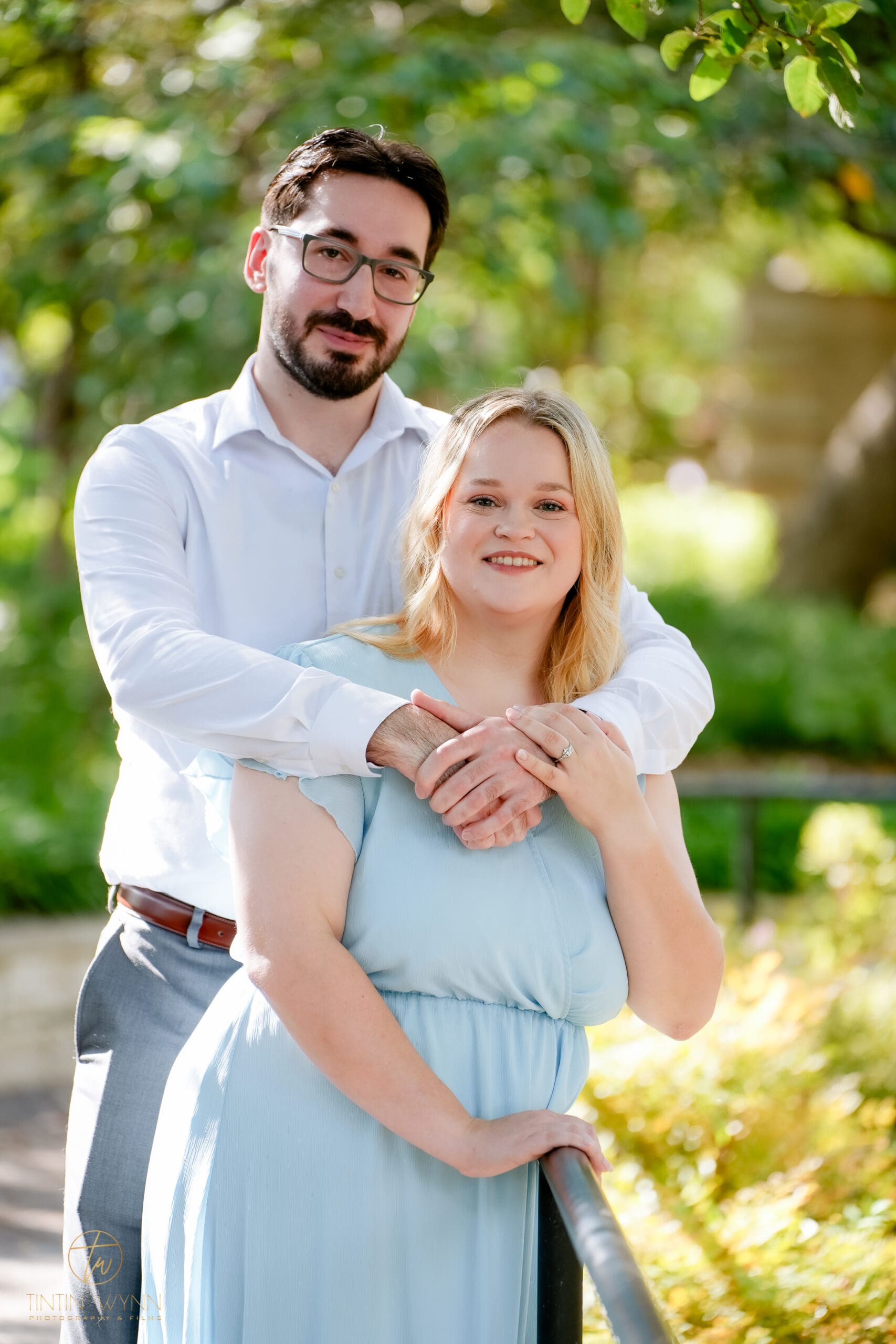 A couple smiles at the camera beside a railing, with arms around each other and a ring visible on their joined hands.