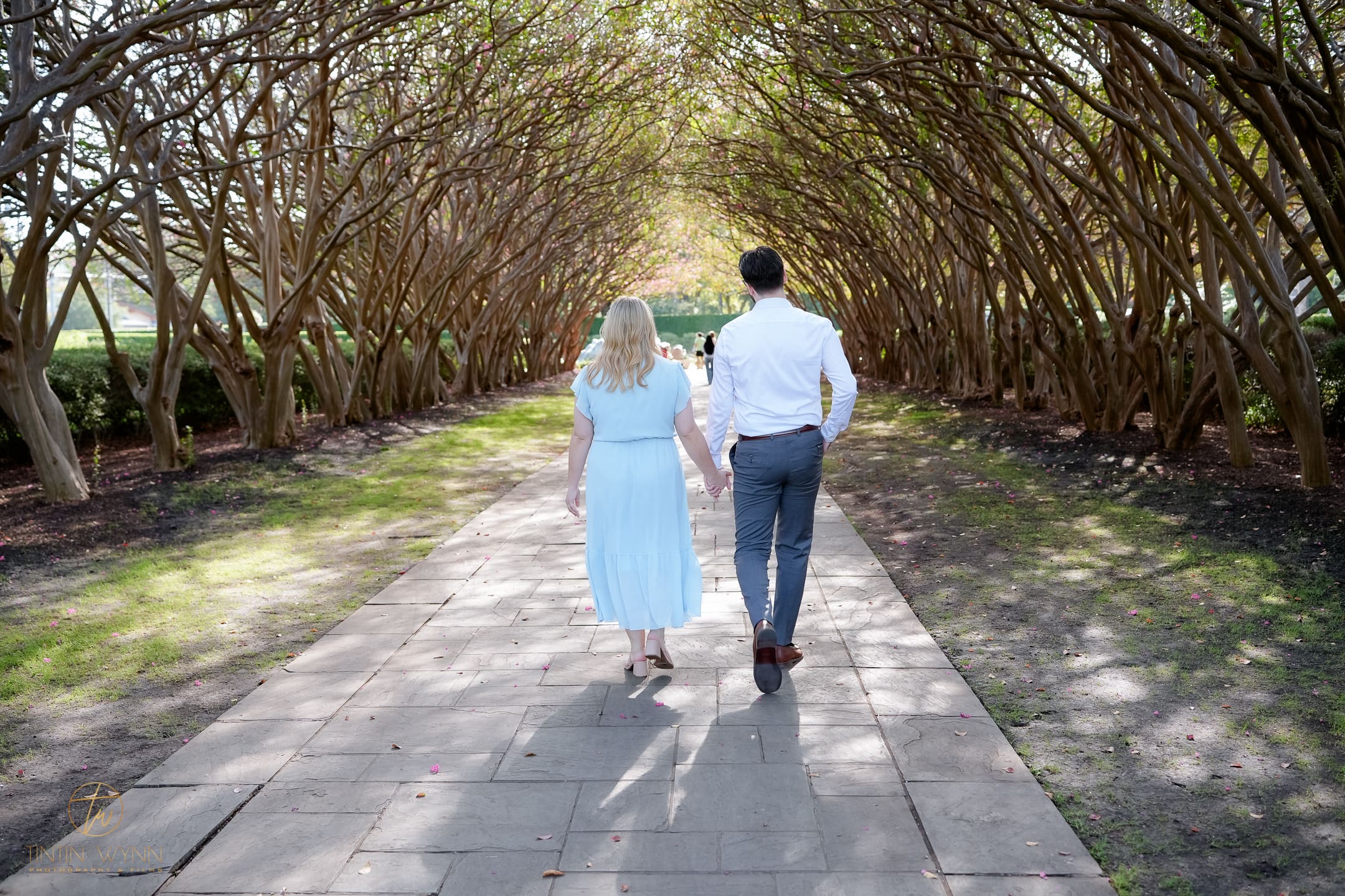 A couple holds hands while walking away along a stone path beneath arching trees.