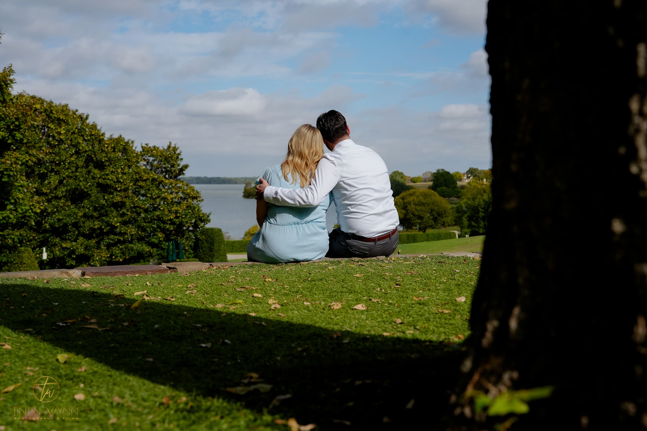 A couple sits together on a stone edge overlooking a lake, framed by a shaded tree and lawn.
