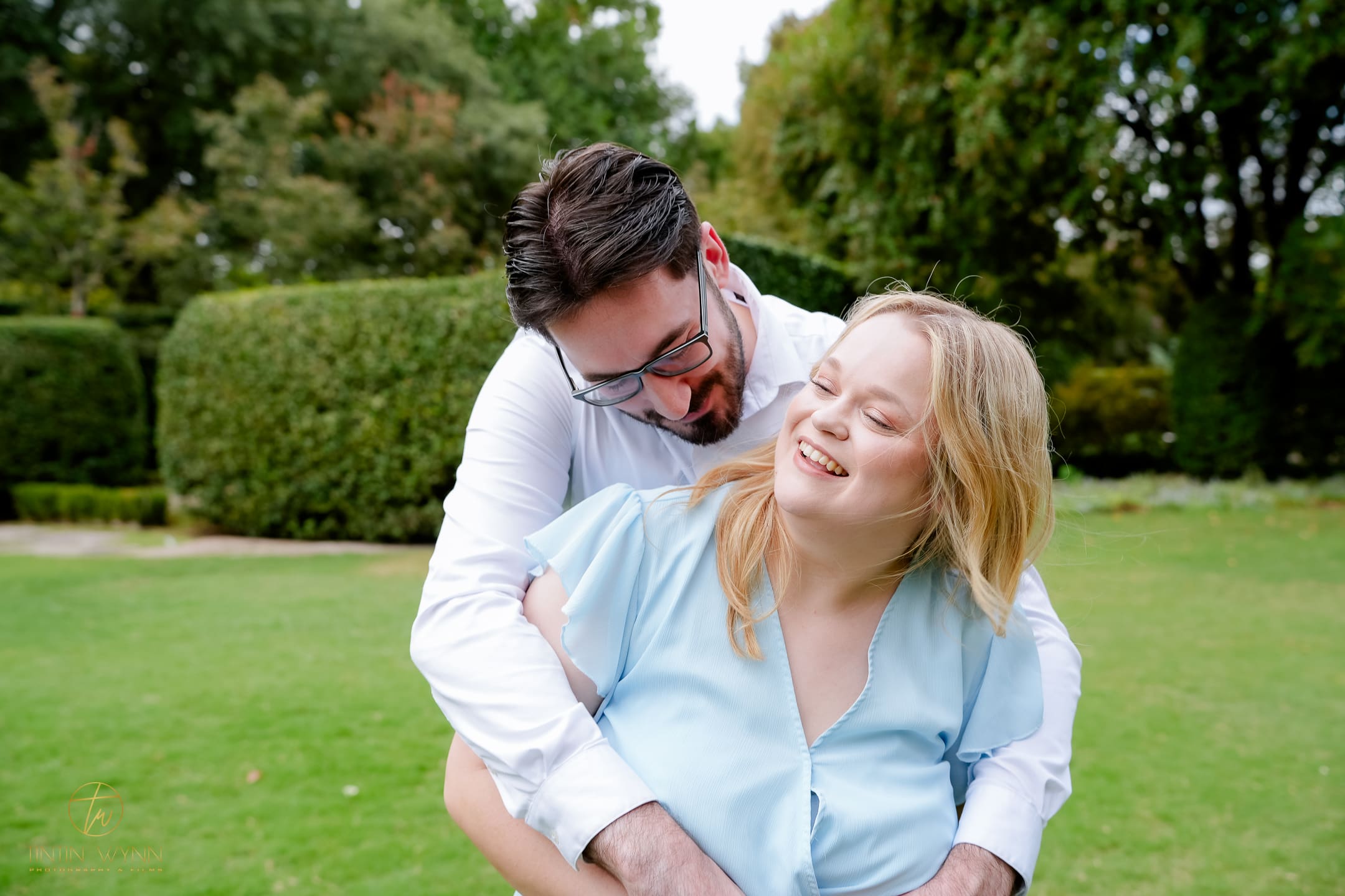 A couple laughs together in a back-hug on a lawn bordered by green hedges.