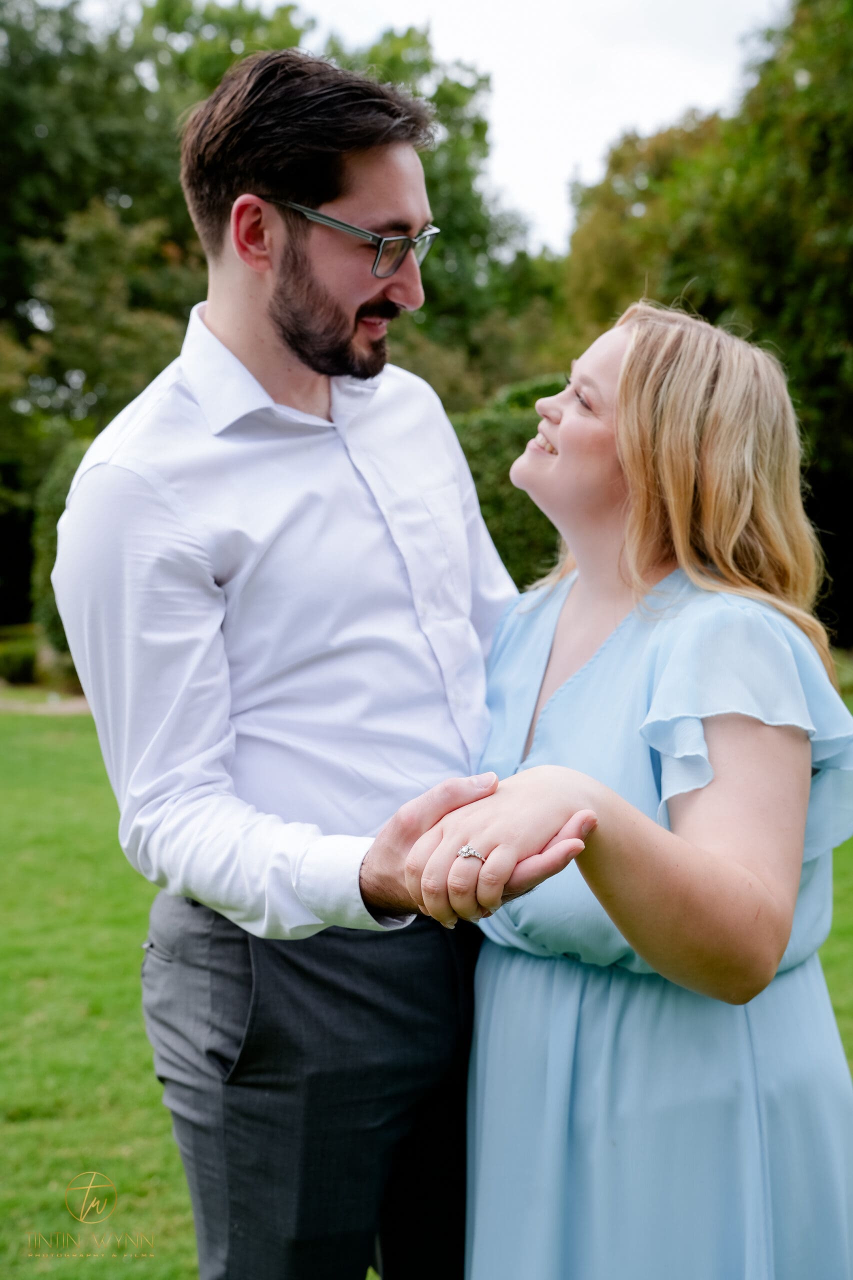 A couple smiles at each other while holding hands, with a ring prominent in the foreground.