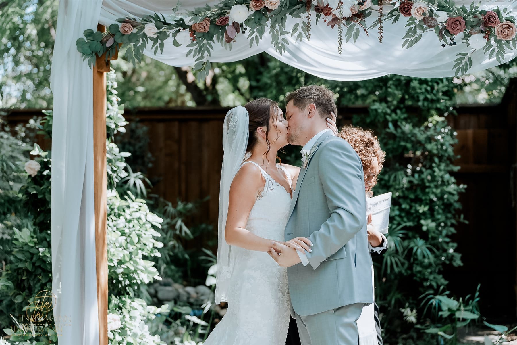 Couple kissing beneath a floral arch draped in white fabric.